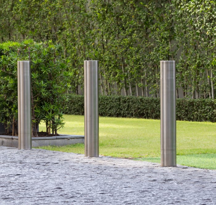 Concrete path in front of a lawn and trees with three silver bollards along the edge of the path.