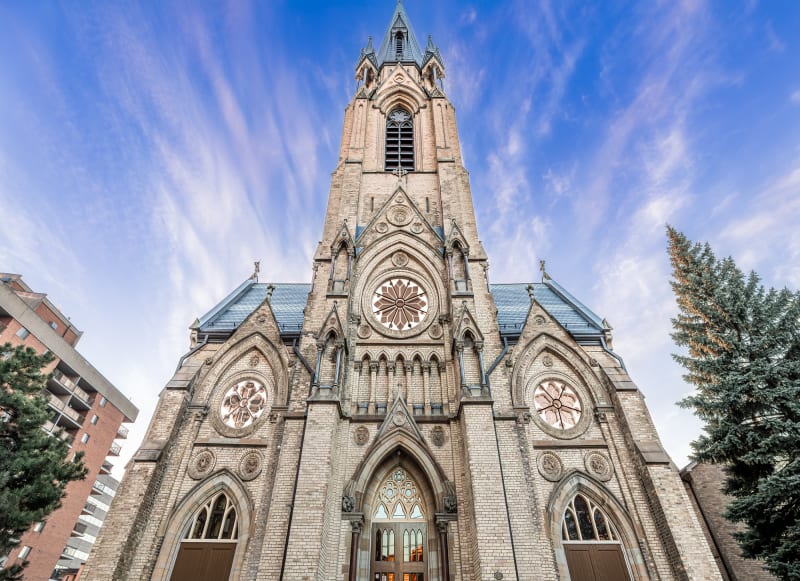 Exterior of a detailed grey brick church with multiple windows and bright blue sky above.