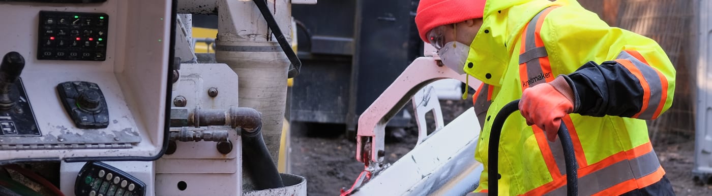 Man in a bright yellow safety jacket, orange hat and gloves, safety glasses, and a white mask, operating concrete machinery.