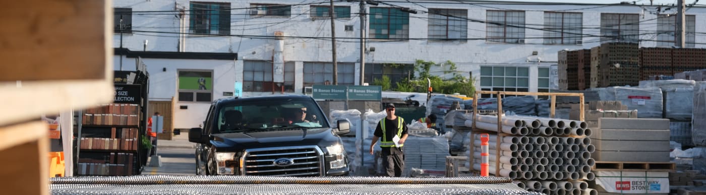 A black pick up truck driving through a construction supply yard with a man in a safety vest walking to the right of the truck.