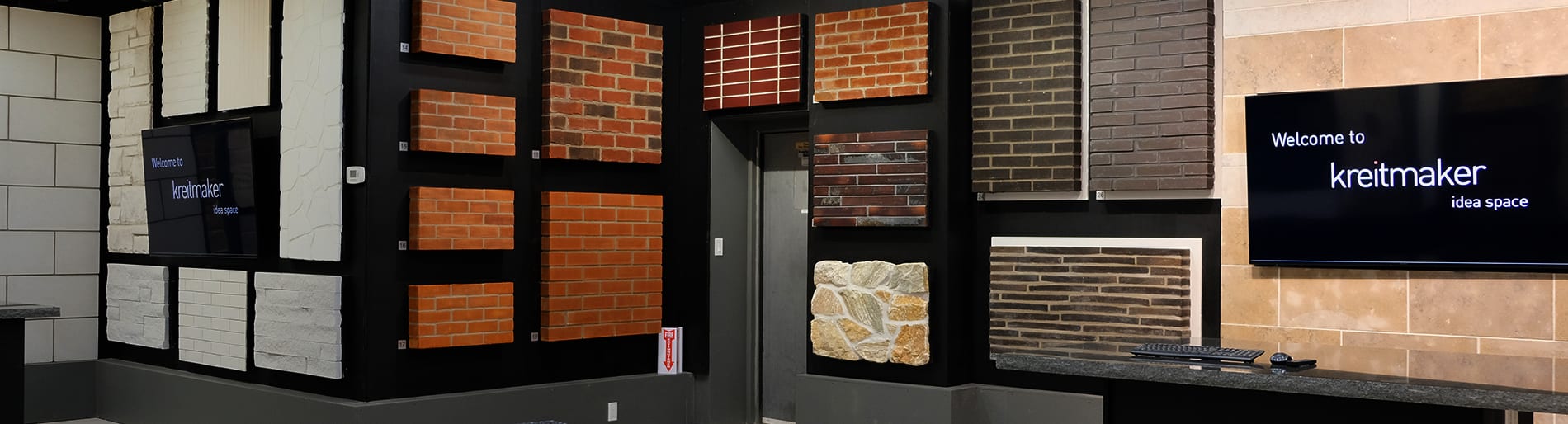 Show room with multiple brick samples in different colours and sizes on the wall and two TV screens, both displaying the [SITE][COMPANY-NAME] logo.