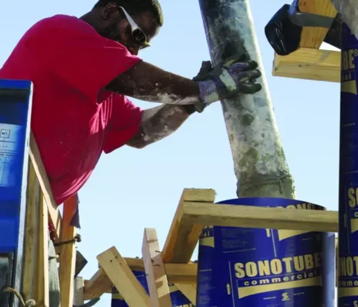 A black man wearing a red t-shirt pouring concrete from a machine into a blue tube.