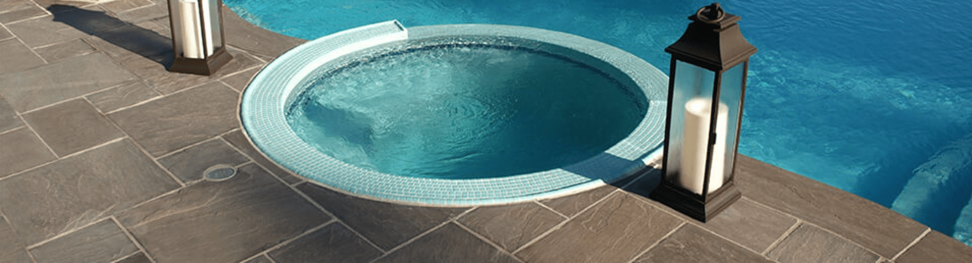 Round hot tub on the edge of a pool with a lantern on either side.