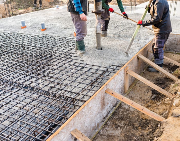 Three men from the shoulders down spreading stone in a wooden frame with a steel rebar grate on top.