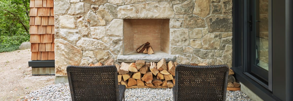 Outdoor beige wall stone fireplace with wood stacked in front of it and chairs facing the fireplace. 