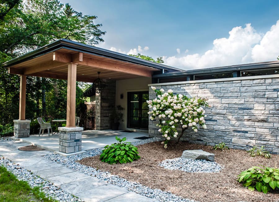 Exterior of a house with light grey wall stone, grey patio stone walkway and wood awning with pillars.